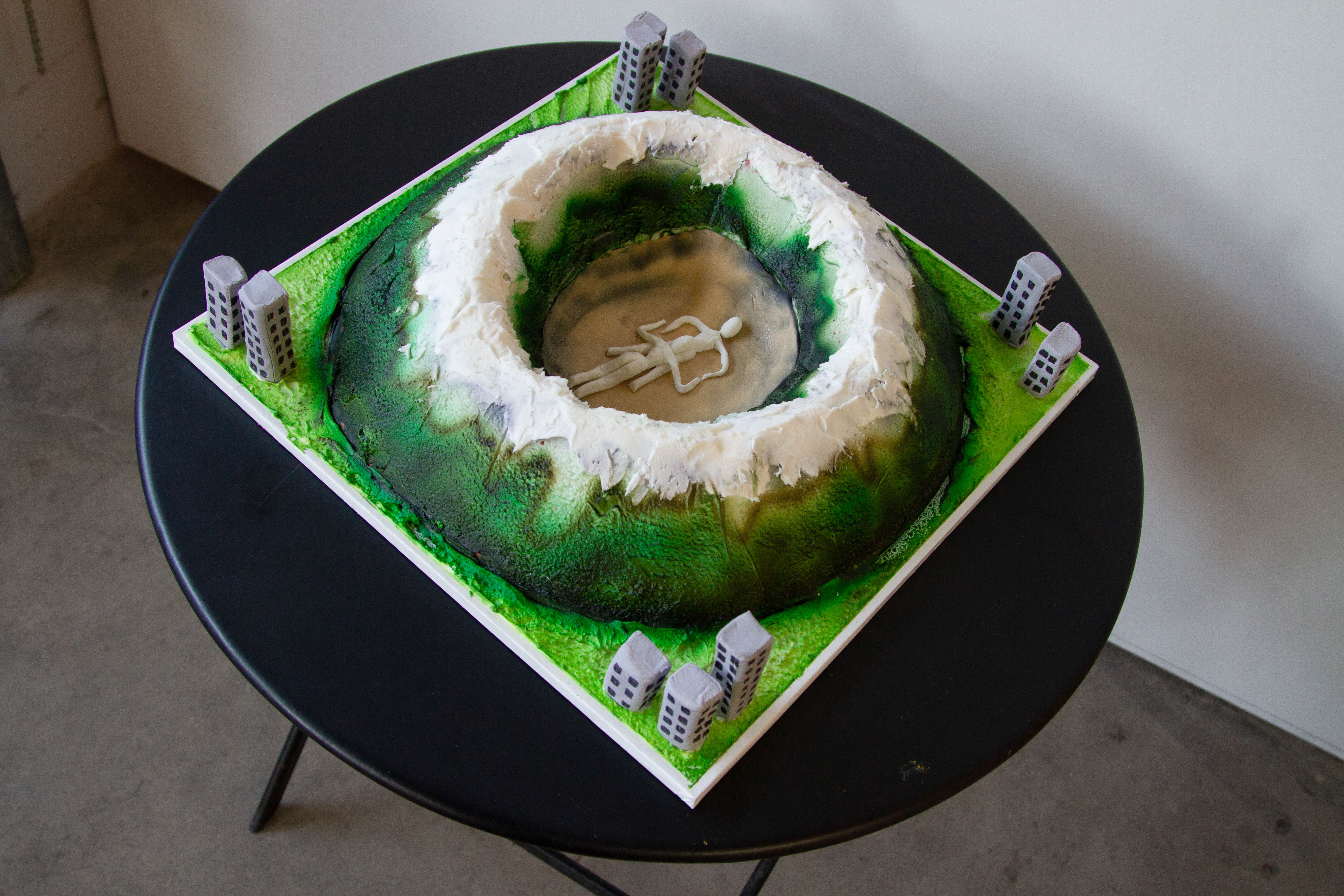 Ariel view of a cake shaped like a mountainous crater with two figures engaged in felatio in the center surrounded by buildings on a black table